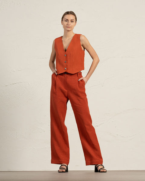 A woman models a cinnamon red linen sleeveless vest and wide-leg pants, standing with hands in pockets against a textured light beige wall. She wears black sandals.