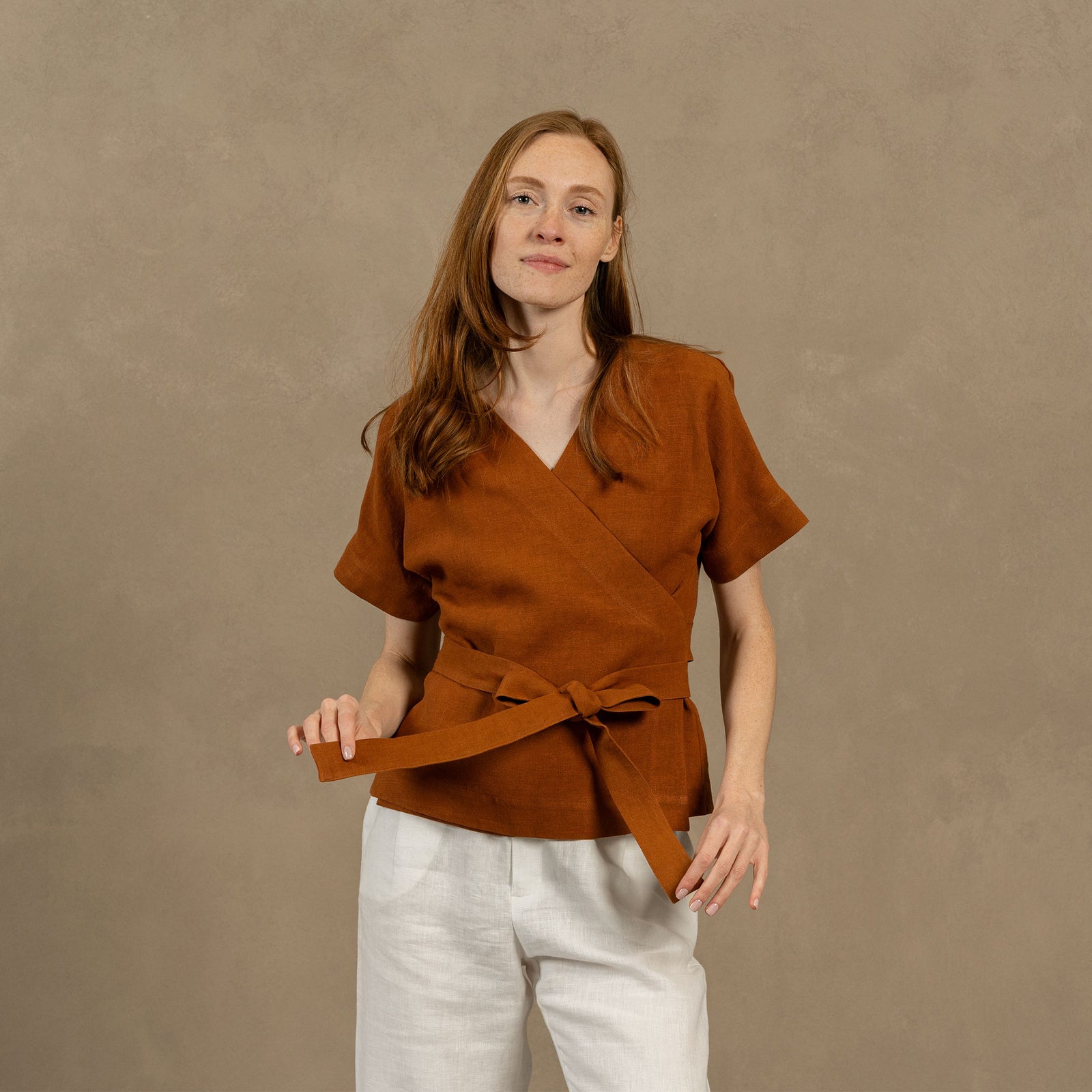A person with light skin and dark hair stands in profile, looking down towards their hands which are crossed at their midsection. They are wearing a linen wrap top with short, loose sleeves and stone green wide-leg pants. In the background, there is a dark metal fence and lush green foliage.