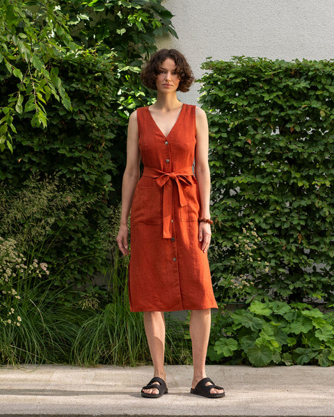 A fair-skinned woman with short, curly dark brown hair stands facing forward, outdoors, wearing a cinnamon red linen dress. The dress is knee-length, sleeveless with a V-neck, and has a row of dark buttons down the front. A tie-belt of the same fabric cinches her waist, tied in a bow. She is also wearing dark, flat sandals. 