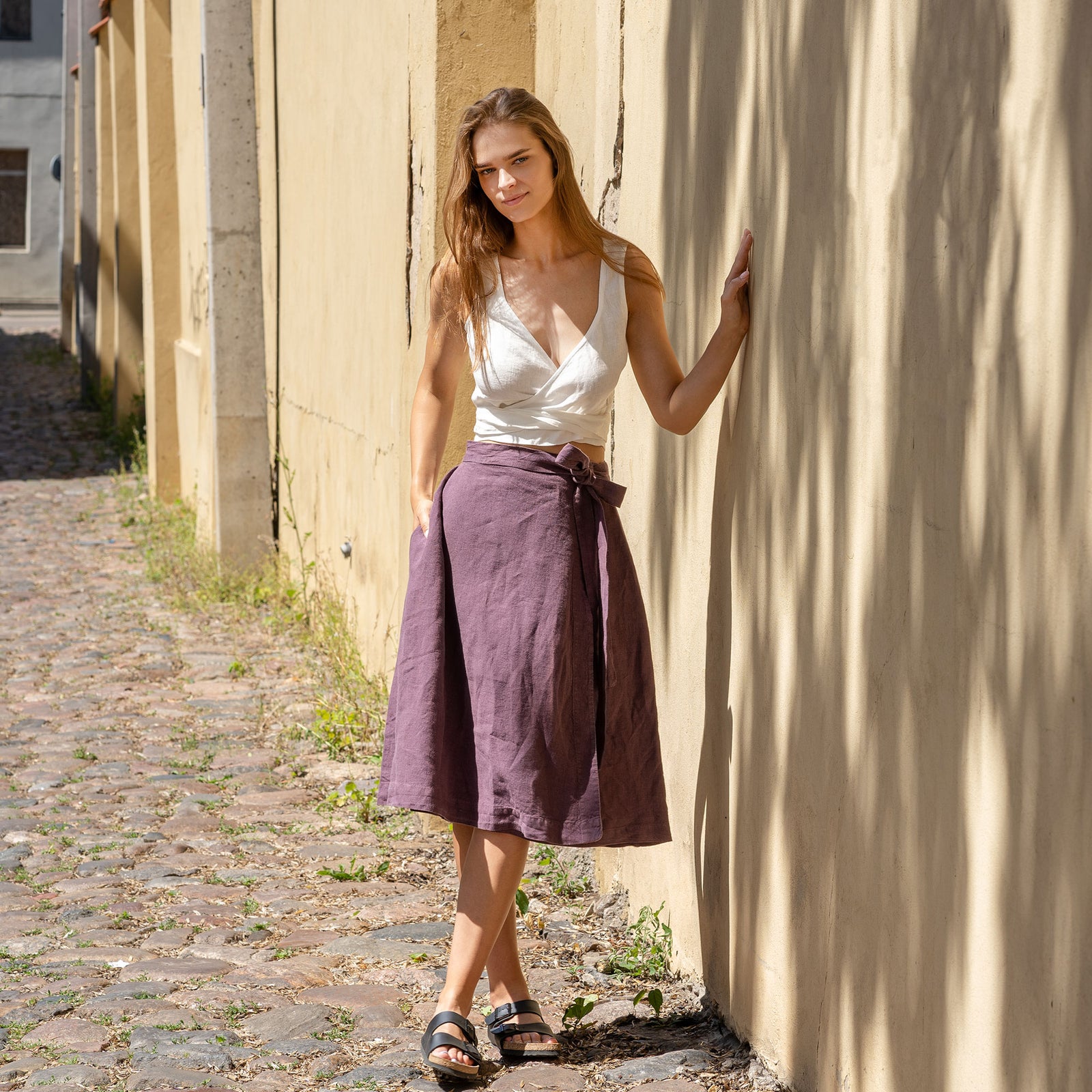 A barefoot woman stands playfully on a stone garden path, lifting one foot slightly off the ground. She is wearing a shadow purple linen wrap skirt, paired with a matching crop top that ties at the waist, surrounded by lush green foliage.