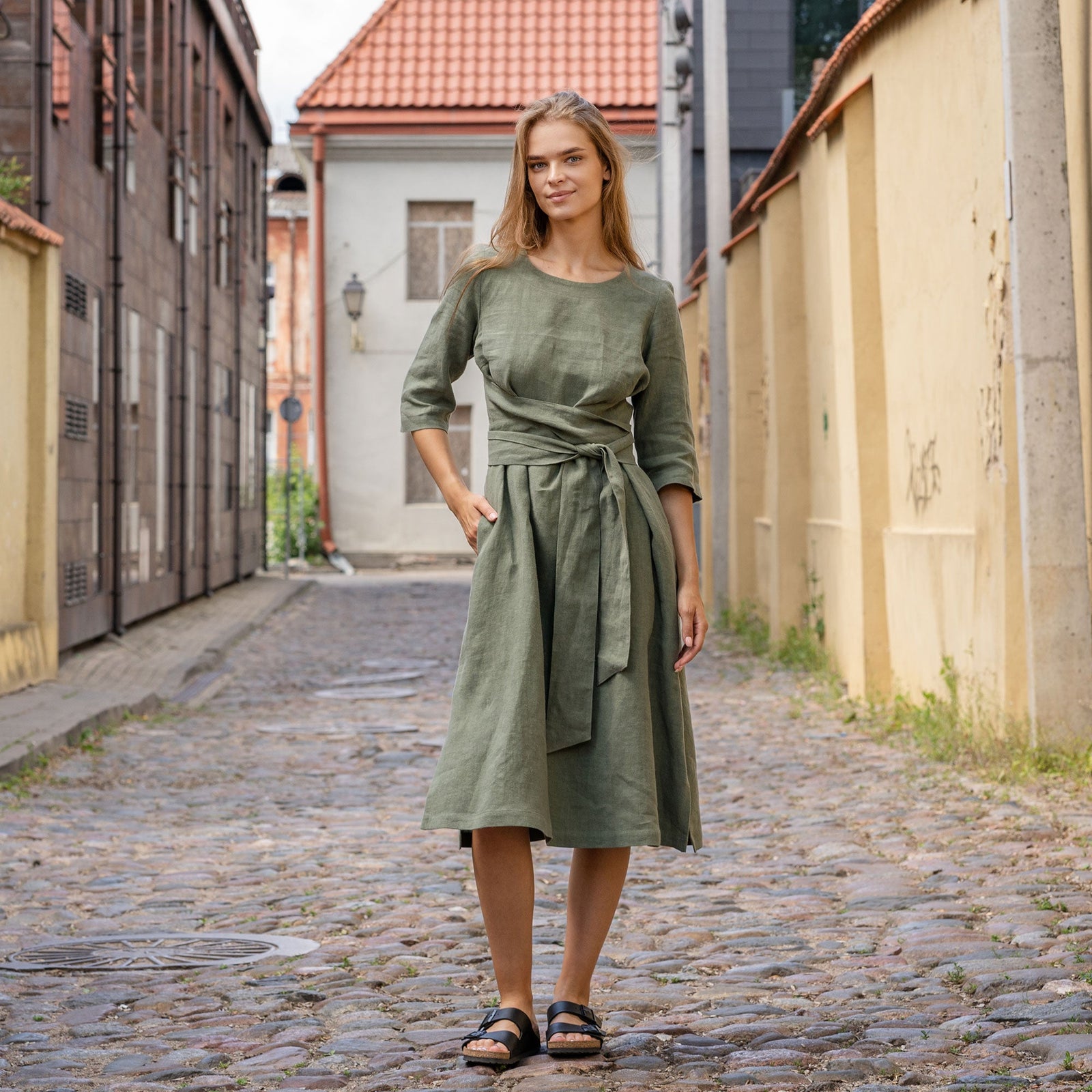 A woman sits on a wooden bench in a park wearing a green dress with her legs elegantly crossed. She holds both hands up near her face while looking slightly upward into the distance.