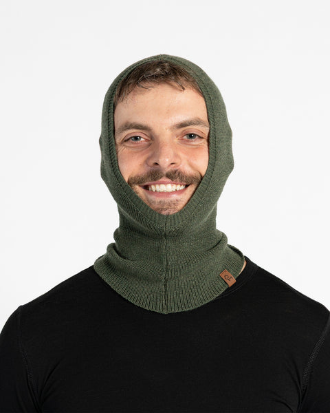 A man with a beard and mustache wears a dark green, fitted menique balaclava that covers his head and neck, with an opening for his face. He is also wearing a black long-sleeved shirt and is looking directly at the viewer with a smile.