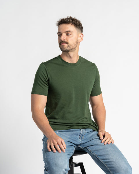 Man in a menique dark green t-shirt and light jeans is seated on a black stool, looking slightly to the side, with a white backdrop.