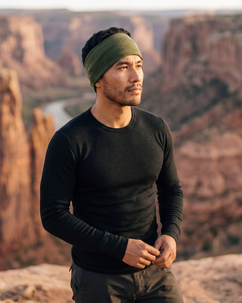A man wearing a black long-sleeve top and an olive green headband stands against a scenic canyon landscape with a river below. He gazes thoughtfully off to the side with his hands casually clasped in front of him.