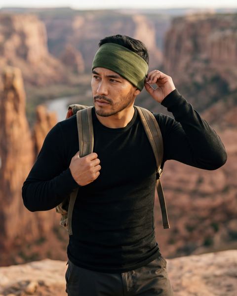 A man wearing a black long-sleeve top, an olive green headband, and a backpack stands on a rocky ledge overlooking a vast canyon and river. He looks off to the side, adjusting his headband with one hand while holding his backpack strap with the other.