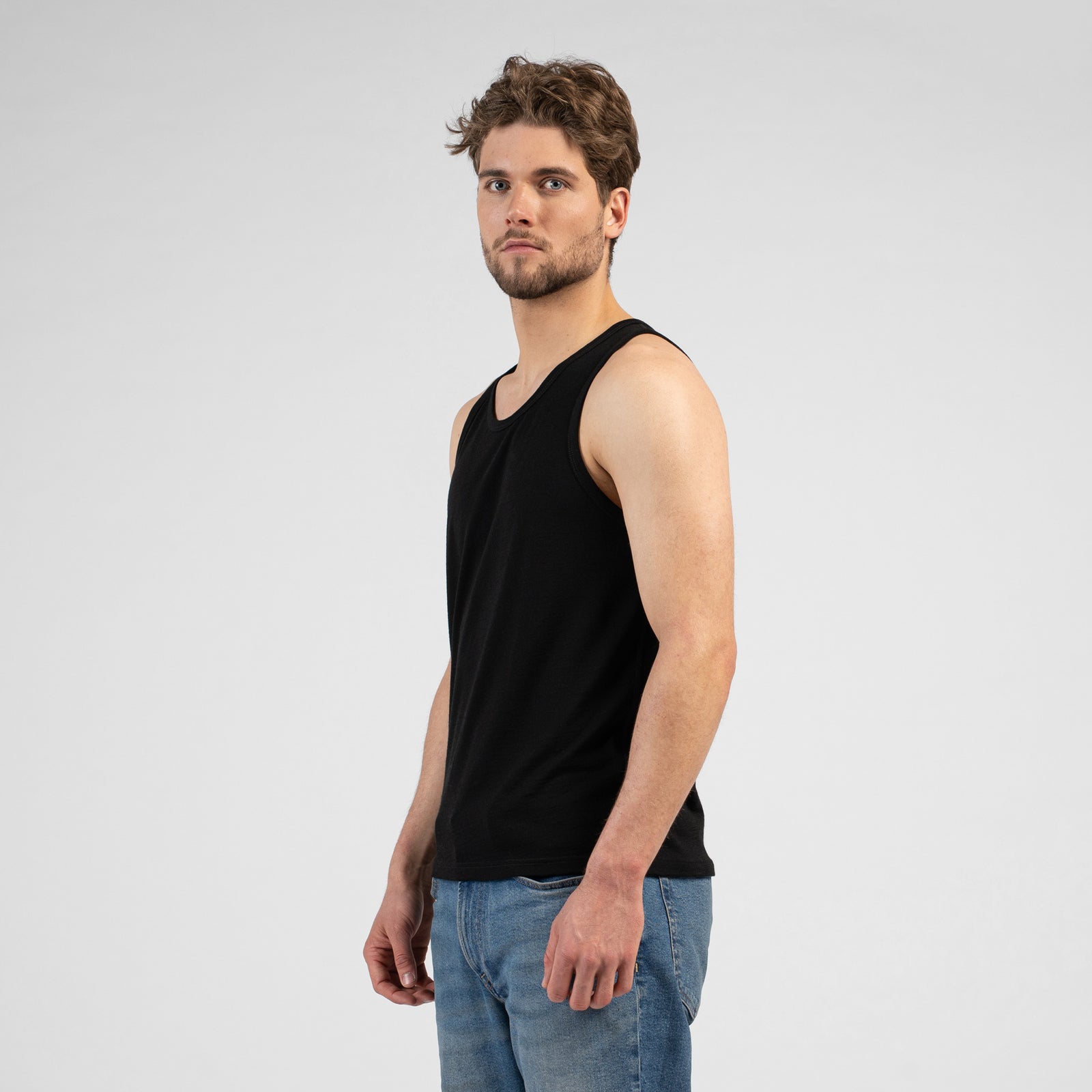 A studio portrait of a man looking off to the left side of the frame with his arms relaxed by his sides. He is wearing a black tank top and blue jeans against a light grey background.