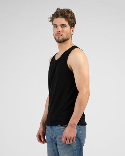 A side-profile studio portrait of a man turning his head to look directly at the camera. He is wearing a black tank top and blue jeans against a plain light grey background.