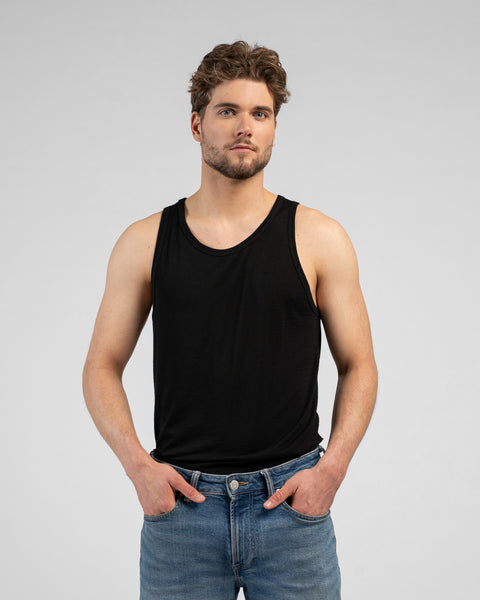 A studio portrait of a man looking directly at the camera with his hands resting in his front pockets. He is wearing a black tank top and blue jeans against a light grey background.