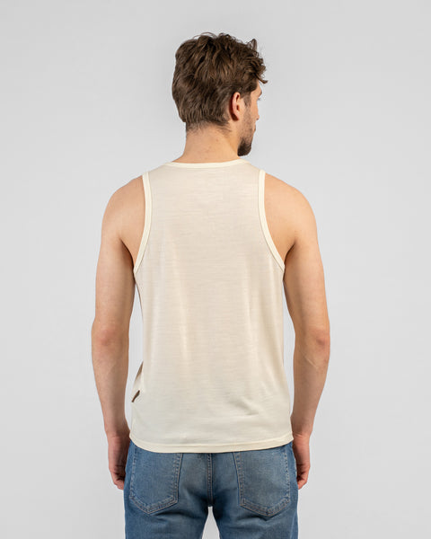 A studio portrait showing the back view of a man wearing a regular fit, light beige tank top and medium-wash blue jeans. The background is a solid light grey.