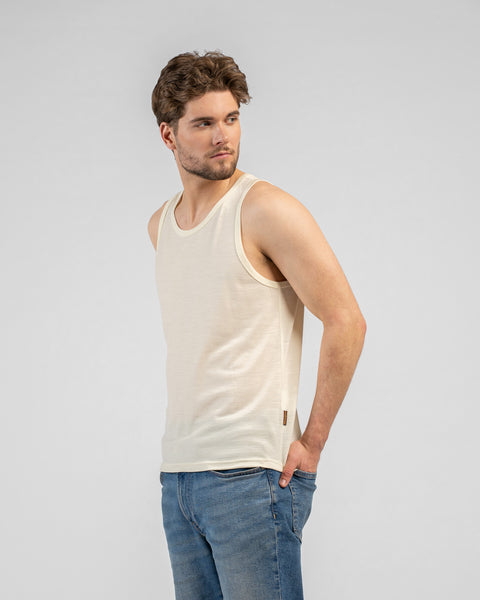 A studio portrait of a man standing and looking back over his right shoulder. He is wearing a light beige tank top and medium-wash blue jeans, with his left hand resting in his front pocket. The background is a solid light grey.