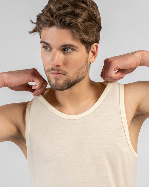 A close-up studio portrait of a man looking off to the left side of the frame. He is wearing a light beige tank top and is holding the shoulder straps with both hands, pulling them slightly outward. The background is a plain light grey.
