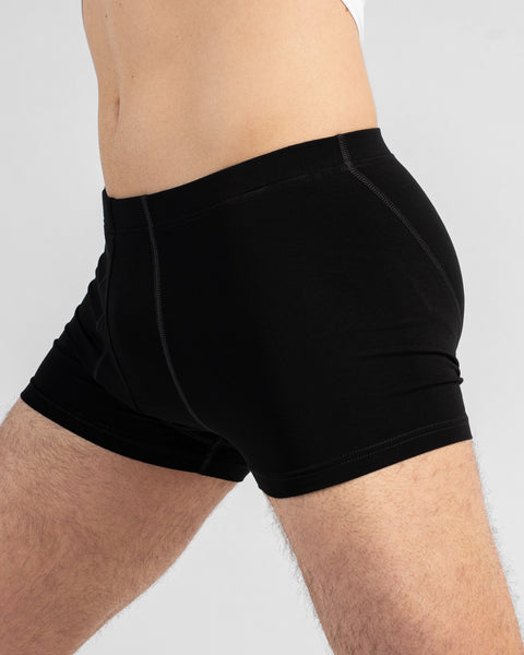 A close-up side profile of a man's torso and upper thighs wearing fitted black boxer briefs. The image displays the waistband, front pouch, and side stitching details against a light grey background.