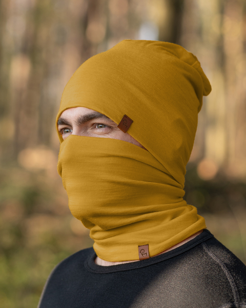 A person with light eyes is wearing a spicy yellow menique beanie that is pulled down to cover part of their forehead and a matching neck gaiter that is pulled up to cover their mouth and nose. They are wearing a dark long-sleeved shirt, and the blurred background suggests an outdoor setting with trees and foliage in muted tones.
