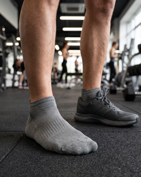 A close-up view of a person's lower legs and feet standing on a black rubber gym floor. The person is wearing gray ankle socks; one foot is shown just in the sock, while the other is wearing a dark gray athletic shoe. The background shows a blurred gym environment.