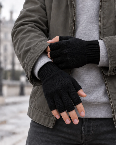 A close-up of a person's hands crossed slightly in front of them, wearing black knit fingerless gloves with ribbed cuffs. The person is dressed in a gray long-sleeved top under an olive green jacket, set against a blurred, overcast outdoor background.