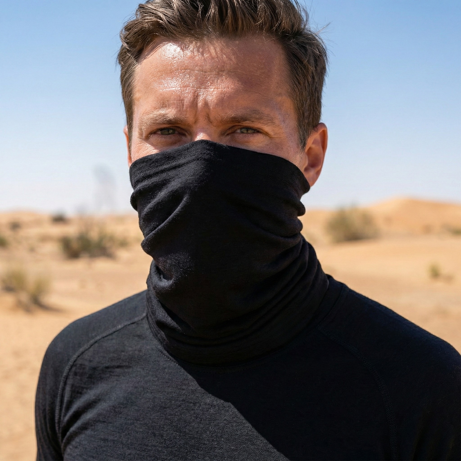 A man outdoors in a wooded area wearing a navy blue long-sleeved hoodie. He has a denim-blue neck gaiter pulled up to cover his lower face and nose. He is grasping the straps of his black backpack and looking back over his shoulder directly at the camera.