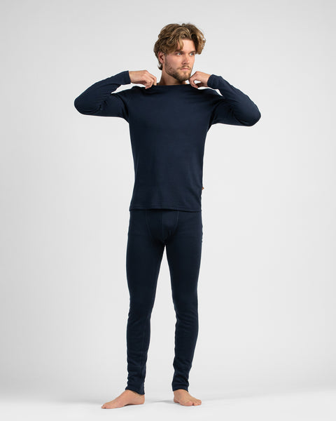 A full-length studio portrait of a barefoot man standing against a white background, wearing dark navy thermal base layers while adjusting his collar.