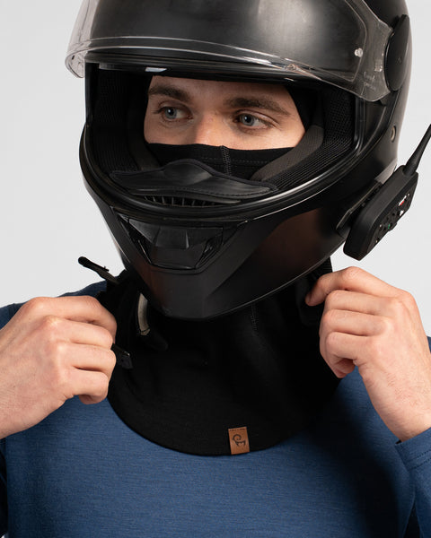 A close-up studio portrait of a man wearing a black balaclava covering his head and lower face, and a blue long-sleeved shirt. He is wearing a black full-face motorcycle helmet with the clear visor up and a communication device attached to the side. He is adjusting the chin strap of the helmet with both hands. The background is a plain light grey.