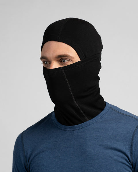 A studio portrait of a man looking off to the left side of the frame. He is wearing a black balaclava that covers his entire head and neck, leaving only his eyes exposed. He is also wearing a blue long-sleeved shirt against a plain light grey background.