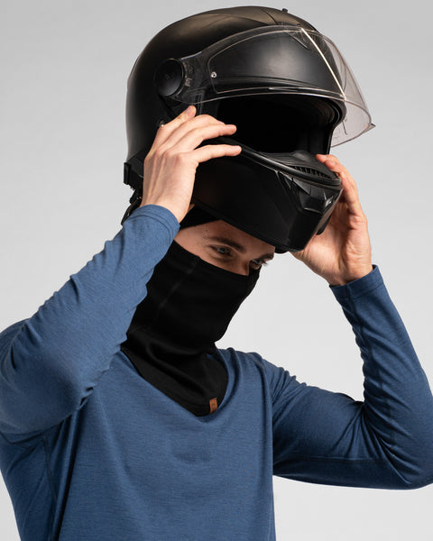 A studio portrait of a man wearing a black balaclava and a blue long-sleeved shirt. He is in the process of putting on a black full-face motorcycle helmet with a clear visor, holding it with both hands as he lowers it onto his head. The background is a solid light grey.