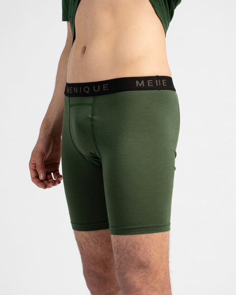 A close-up profile shot of a man wearing dark green long boxer briefs with a black "MENIQUE" waistband. He pulls up a matching dark green t-shirt to show his midriff against a light grey background.