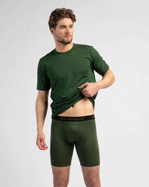 A studio portrait of a man looking to the left while lifting the front hem of his dark green short-sleeved t-shirt. He is wearing matching dark green long boxer briefs with a black logo waistband against a light grey background.