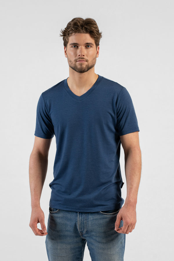 A studio portrait of a man looking directly at the camera with his hands relaxed by his sides. He is wearing a denim blue v-neck t-shirt and blue jeans against a light grey background.