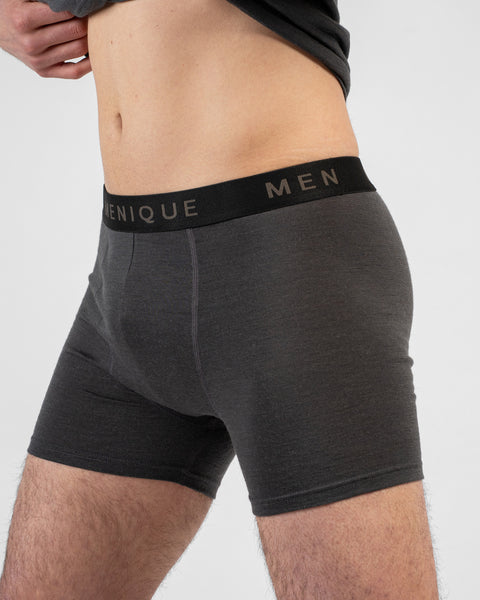 A close-up side profile shot of a man's torso and thighs wearing charcoal grey short boxer briefs with a black "MENIQUE" waistband. He is lifting his dark grey t-shirt to reveal his midriff.