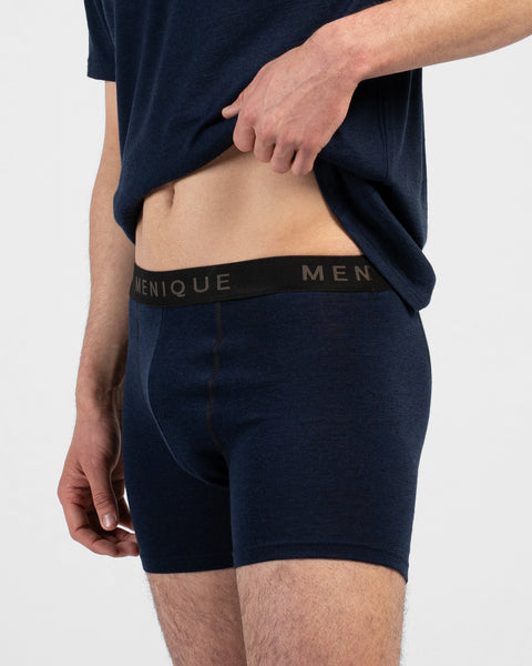 A close-up lifestyle shot of a man's torso and thighs against a plain white background. He is lifting the hem of a dark navy blue t-shirt with his left hand to reveal matching dark navy blue short boxer briefs. The underwear features a contrasting black elastic waistband with "MENIQUE" woven in light brown text.