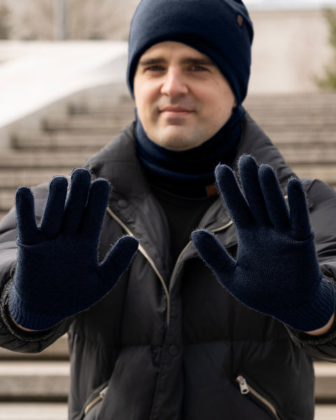 A person wearing a dark blue menique knitted hat, a matching neck gaiter, and navy blue gloves, holding both hands up towards the camera with palms facing forward. They are wearing a dark puffer jacket. The background is blurred, showing concrete steps.