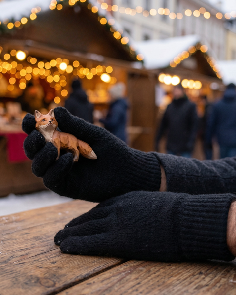 A close-up of a person's hands resting on a snow-dusted wooden table at an outdoor holiday market. The person is wearing thick, black knitted wool gloves and holding a small, carved wooden fox figurine in one hand. The background is filled with blurred, warmly lit wooden market stalls and silhouettes of people.
