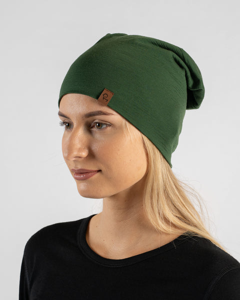 A studio portrait of a blonde woman looking slightly down and to the left against a plain light grey background. She is wearing a black long-sleeved shirt and a dark green slouchy beanie with a small brown logo tag.