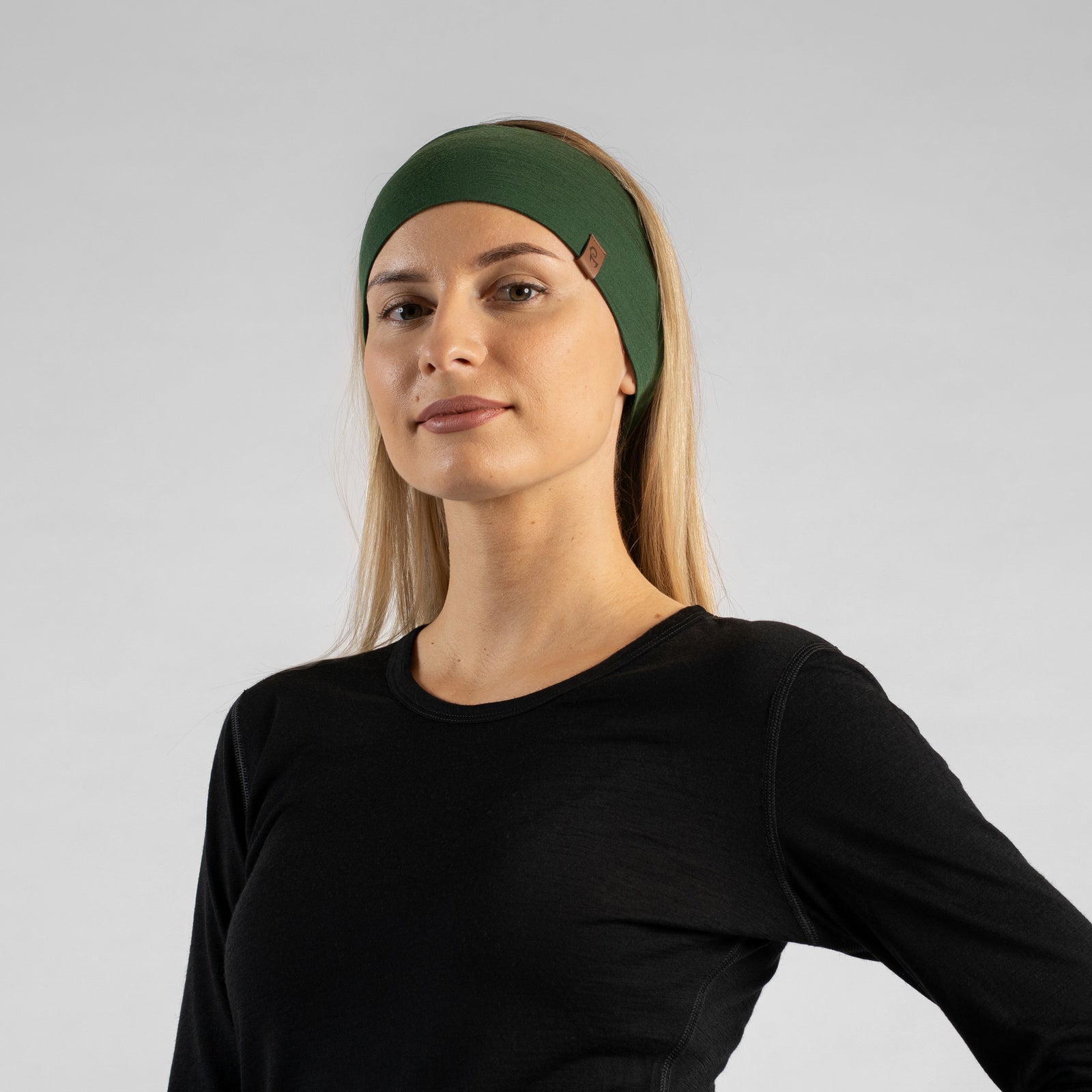 The picture features a woman sitting down in a forest, wearing the unisex dark green menique merino HEADBAND. She is also wearing a sleeveless black merino top. She is looking to the side.