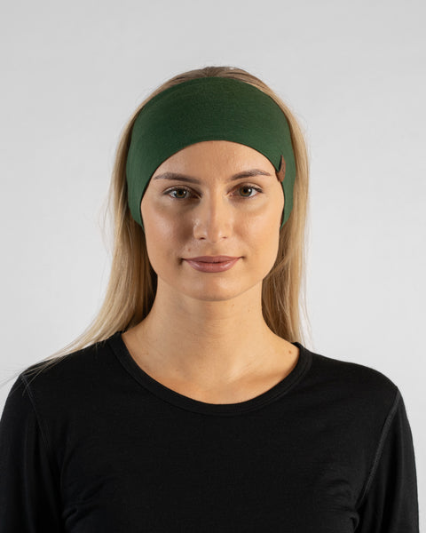 A studio portrait of a blonde woman facing forward and looking directly at the camera. She is wearing a black long-sleeved shirt and a dark green headband against a plain light grey background.