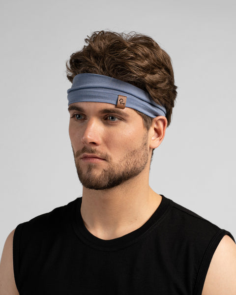A studio portrait of a man looking off to the left side of the frame. He has wavy brown hair and a short beard. He is wearing a dusty blue headband featuring a small brown leather logo patch on the front-left edge. He is wearing a black sleeveless top against a plain light grey background.