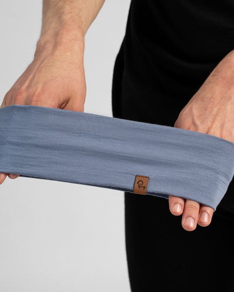 A close-up shot of a person holding a wide, dusty blue headband stretched between their hands. The headband features a small brown logo tag on the bottom edge.