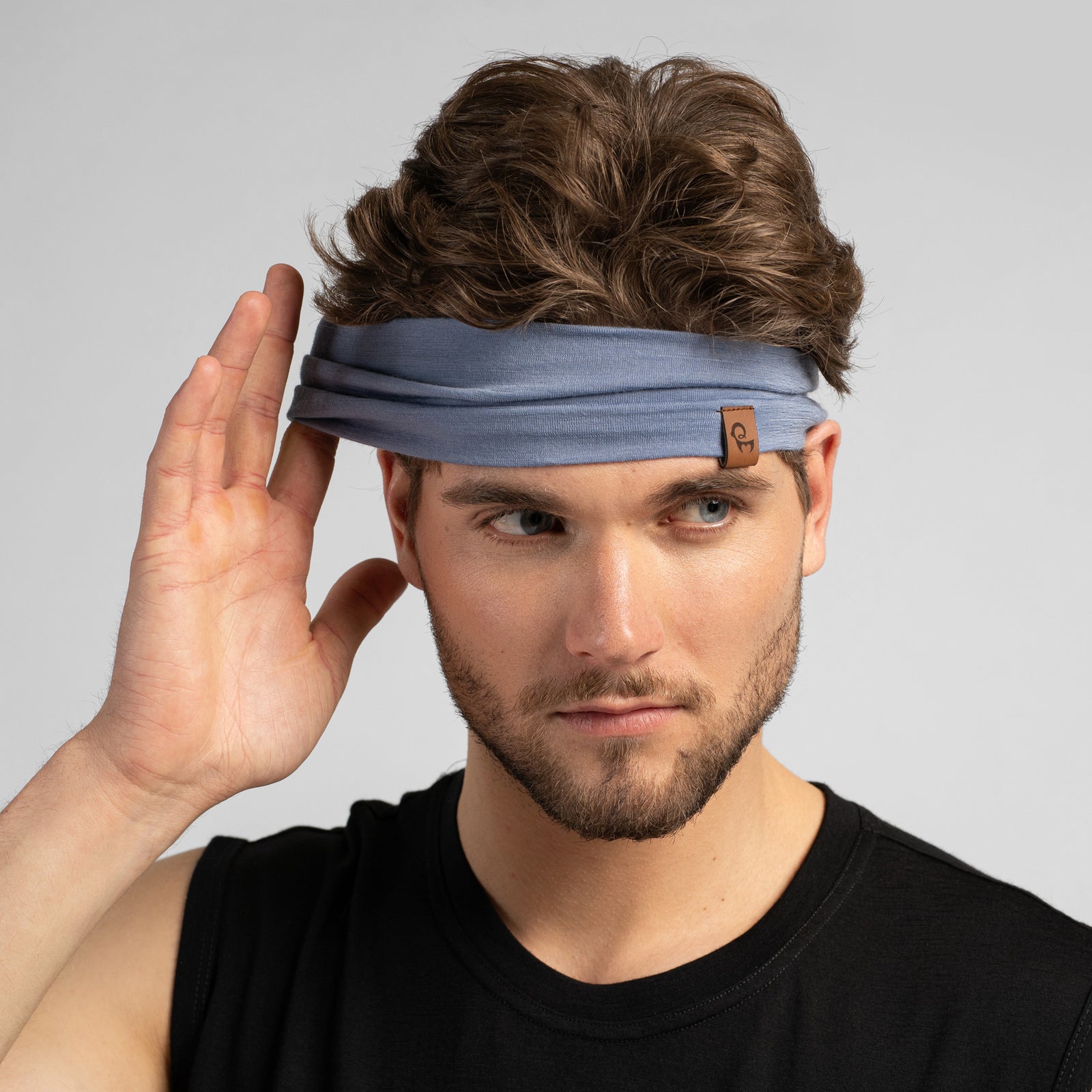 A studio portrait of a man looking off to the left side of the frame. He has wavy brown hair and a short beard. He is wearing a dusty blue headband featuring a small brown leather logo patch on the front-left edge. He is wearing a black sleeveless top against a plain light grey background.