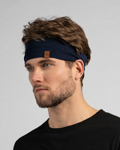 A studio portrait of a man looking off to the left side of the frame. He has wavy brown hair and a short beard. He is wearing a dark navy blue headband featuring a small brown leather logo patch on the front-left edge. He is wearing a black long-sleeved top against a plain light grey background.
