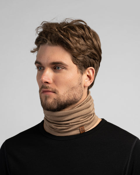 A studio portrait of a man looking off to the left against a plain light grey background. He is wearing a black long-sleeved shirt and a beige neck gaiter gathered around his neck, featuring a small brown logo tag.