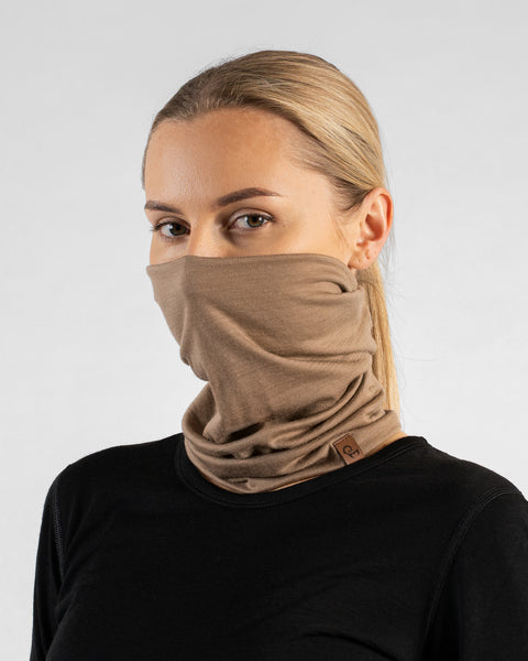 A studio portrait of a blonde woman looking directly at the camera. She is wearing a light beige neck gaiter pulled up to cover her nose and lower face, along with a solid black long-sleeved top. A small brown leather logo patch is visible on the bottom edge of the gaiter. The background is a plain light grey.