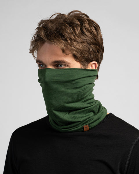 A studio portrait of a man looking off to the left. He is wearing a black long-sleeved shirt and a dark green neck gaiter pulled up to cover his lower face and nose.