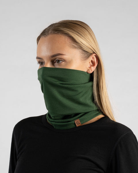 A studio portrait of a blonde woman looking off to the left against a plain light grey background. She is wearing a black long-sleeved shirt and a dark green neck gaiter pulled up to cover her nose and mouth, featuring a small brown logo tag at the bottom.