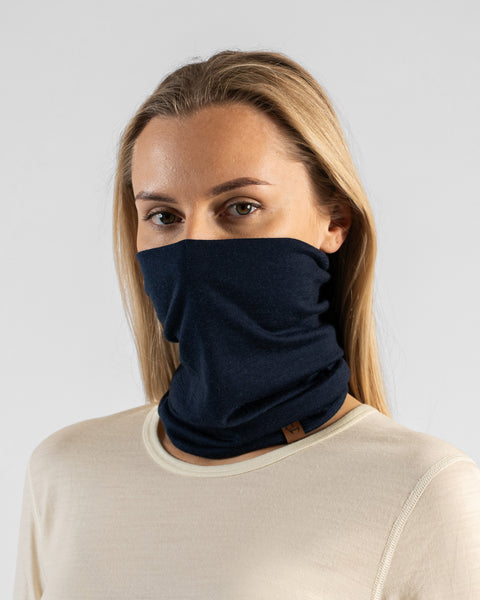 A studio portrait of a blonde woman looking directly at the camera. She is wearing a dark navy blue neck gaiter pulled up over her nose, covering her lower face. The gaiter has a small brown leather logo patch on the bottom edge. She is wearing a light cream, natural-colored long-sleeved top against a plain light grey background.