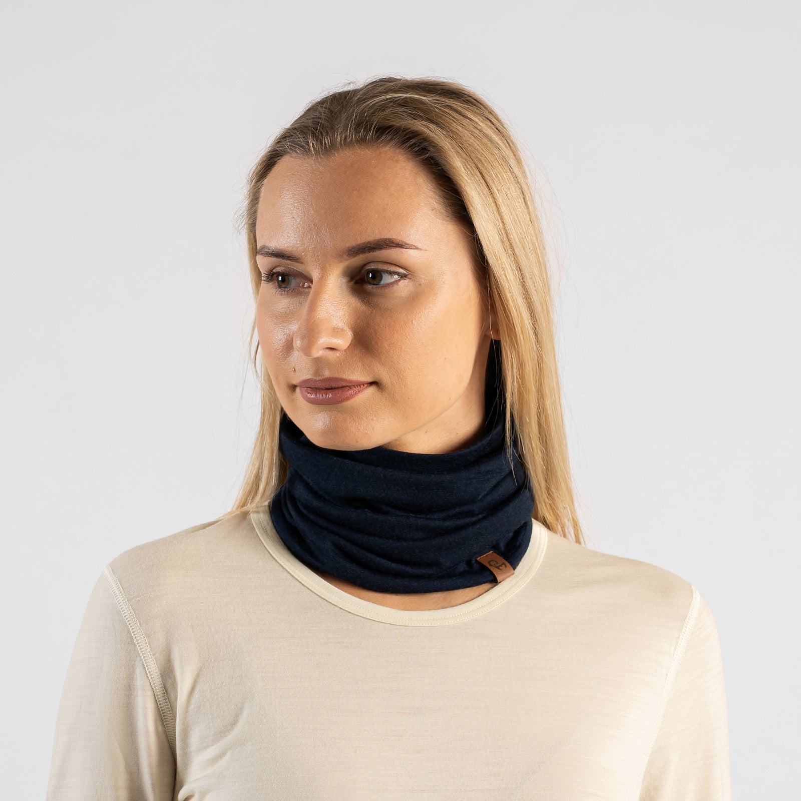 A studio portrait of a blonde woman looking directly at the camera. She is wearing a dark navy blue neck gaiter pulled up over her nose, covering her lower face. The gaiter has a small brown leather logo patch on the bottom edge. She is wearing a light cream, natural-colored long-sleeved top against a plain light grey background.
