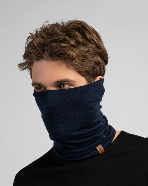 A studio portrait of a man looking thoughtfully off to the left side of the frame. He is wearing a dark navy blue neck gaiter pulled up over his nose, covering his lower face. The gaiter has a small brown leather logo patch on the bottom edge. He is wearing a solid black long-sleeved top against a plain light grey background.