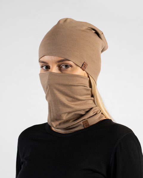 A studio portrait of a blonde woman looking directly at the camera. She is wearing both a light beige slouchy beanie and a matching light beige neck gaiter pulled up to cover her nose and lower face. Both items feature a small brown leather logo patch. She is wearing a solid black long-sleeved top against a plain light grey background.