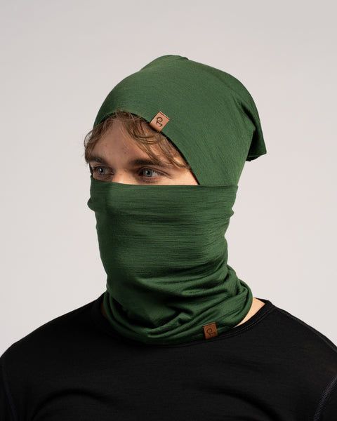 A studio portrait of a man looking to the left against a plain light grey background. He is wearing a black shirt, a dark green slouchy beanie, and a matching dark green neck gaiter pulled up over his nose and mouth.