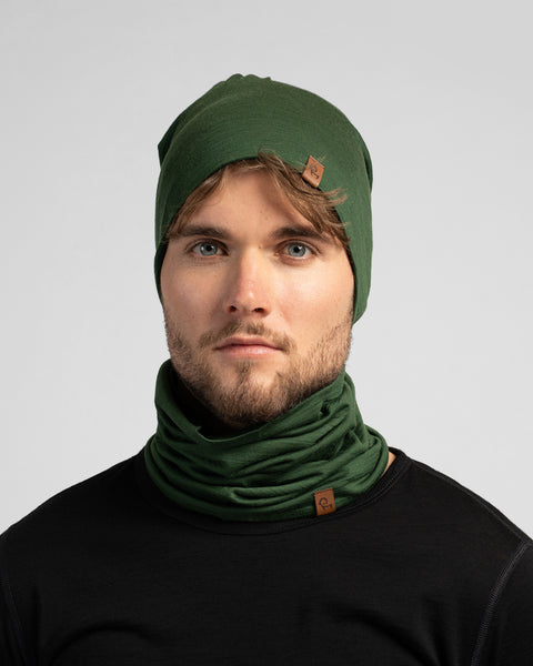 A studio portrait of a man looking directly at the camera. He is wearing a black long-sleeved shirt and a matching set of a dark green slouchy beanie and neck gaiter. Both the beanie and gaiter feature a small brown logo tag.