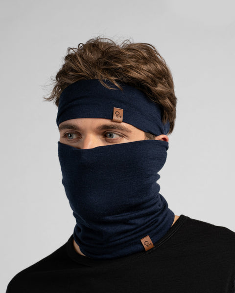 A studio portrait of a man looking thoughtfully off to the left side of the frame. He is wearing a dark navy blue neck gaiter pulled up over his nose, covering his lower face, along with a matching dark navy blue headband. Both items feature a small, square brown leather logo patch. He is wearing a solid black long-sleeved top against a plain light grey background.