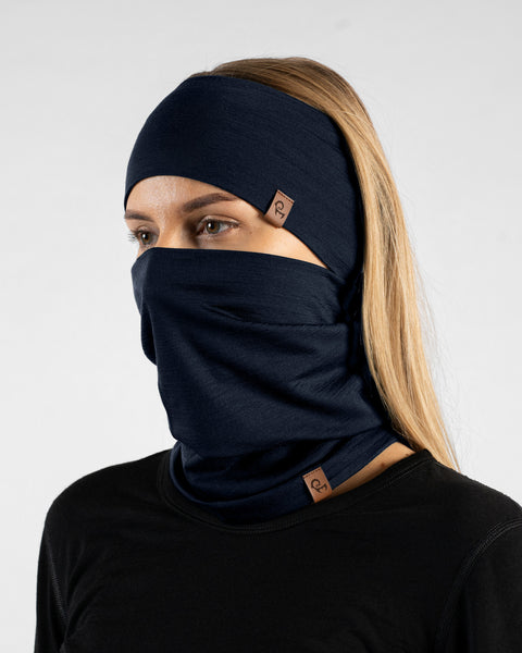 A studio portrait of a blonde woman looking thoughtfully off to the left side of the frame. She is wearing a dark navy blue neck gaiter pulled up over her nose, covering her lower face, along with a matching dark navy blue headband. Both items feature a small, square brown leather logo patch. She is wearing a solid black long-sleeved top against a plain light grey background.